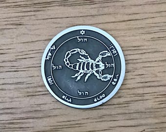 May include: A circular silver-toned metal coin featuring a raised scorpion graphic in the center. The coin has a dark grey recessed background and a border inscribed with Hebrew characters and a small Star of David symbol at the top position.