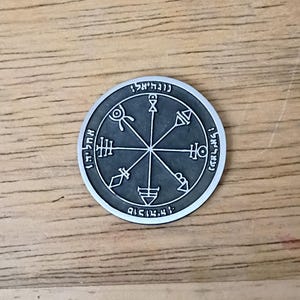 May include: A circular silver-toned metal coin featuring a dark gray recessed center. The surface displays eight radiating lines ending in distinct geometric symbols, surrounded by a border inscribed with Hebrew characters in a raised silver finish.