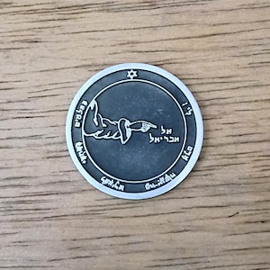 May include: A circular silver-toned metal coin featuring a raised relief of a pointing hand in the center. The design includes a small Star of David at the top and Hebrew text engraved along the inner border and near the hand, with a dark finish.