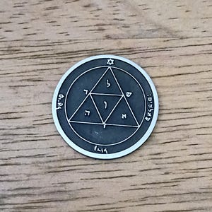 May include: A circular silver-toned metal medallion featuring a dark recessed background. The design includes a central triangle containing Hebrew characters, surrounded by a larger circle with a small Star of David and additional Hebrew text.