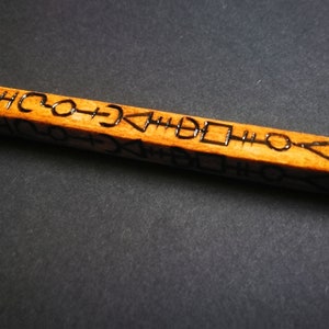 May include: A wooden wand with a brown finish and black markings. The markings are a series of symbols and shapes.