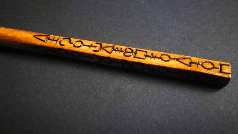 May include: A wooden wand with a brown finish and black markings. The markings are a series of symbols and shapes.
