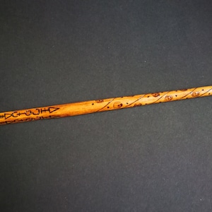 May include: A wooden wand with a brown finish and intricate carvings. The wand is decorated with a repeating pattern of geometric shapes and circles.