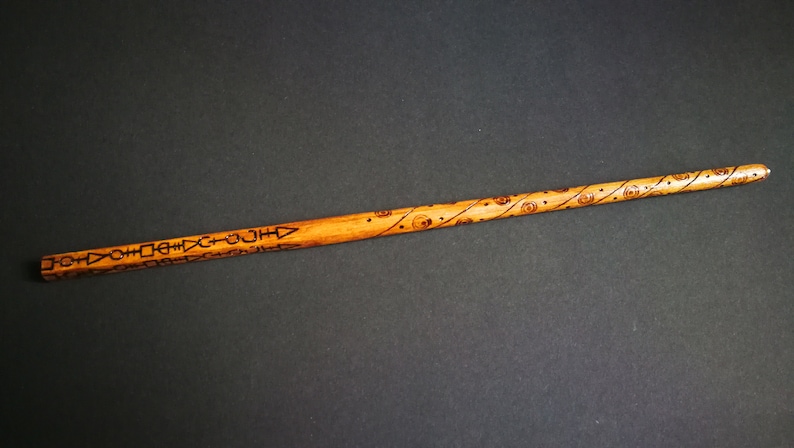 May include: A wooden wand with a brown finish and intricate carvings. The wand is decorated with a repeating pattern of geometric shapes and circles.