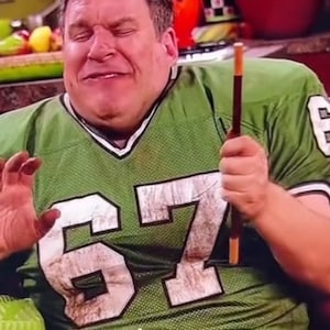 May include: A man wearing a green and white football jersey with the number 67 on the front. He is holding a brown wooden stick in his right hand and has a serious expression on his face.