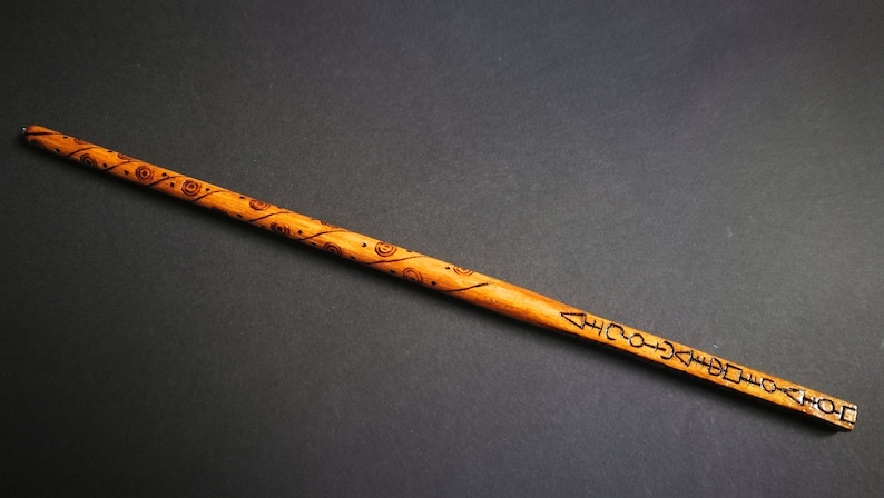 May include: A wooden wand with a brown finish and intricate carvings. The wand is decorated with a swirling pattern and symbols.