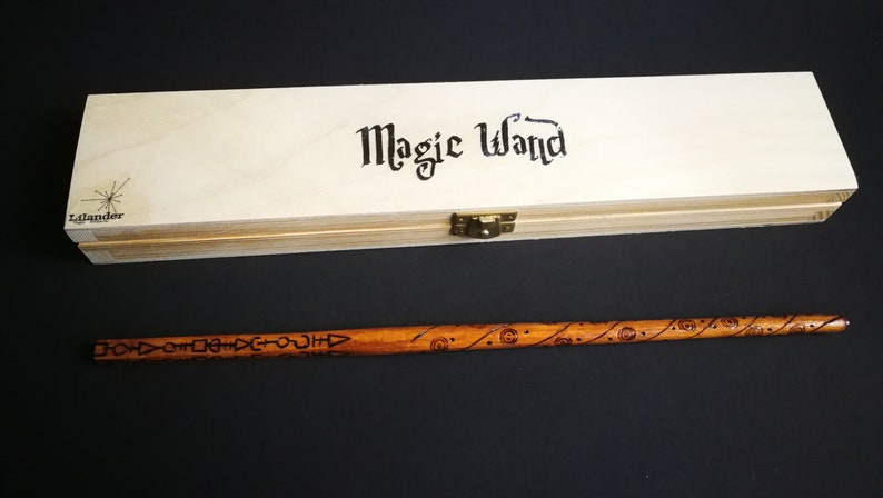 May include: A wooden box with the words "Magic Wand" written on the top. The box is closed and has a brass latch. A wooden wand with intricate carvings is lying next to the box.
