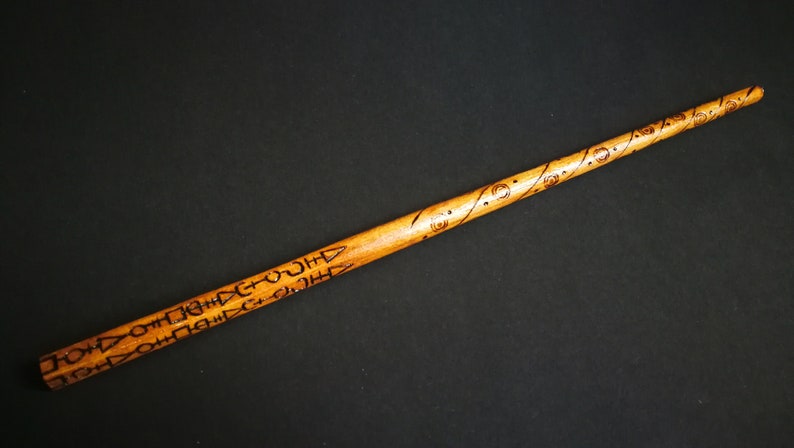 May include: A wooden wand with intricate carvings and a dark brown finish. The wand is approximately 12 inches long.