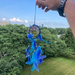 May include: A blue dreamcatcher with a tree of life design. The dreamcatcher features a blue hoop, adorned with purple stones and blue feathers. The dreamcatcher is suspended by a thin blue string.