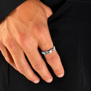 Op de afbeelding: Een zilverkleurige metalen ring met een golvend, golvend bandontwerp. De ring heeft een gladde, gepolijste afwerking en een doorlopende, onregelmatige rand die een vloeiende, organische vorm rond de vinger creëert, wat een eenvoudige metalen uitstraling geeft.