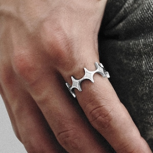 Könnte beinhalten: Ein silberner Ring mit einem sich wiederholenden Sternmuster. Der Ring wird an einem Finger getragen, und das Design umschließt den Finger. Der Ring besteht aus glänzendem Metall und hat eine moderne, geometrische Ästhetik.
