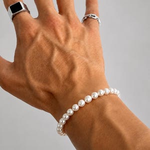 May include: A hand displaying a collection of jewelry. The hand wears a silver ring with a black square stone, a chain-link ring, and a pearl bracelet. The jewelry is set against a neutral background.