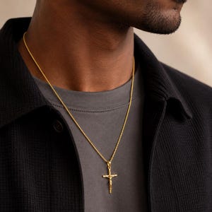 May include: A gold-toned metal crucifix pendant suspended on a thin, textured rope chain. The pendant features a detailed figure of a person on a cross, crafted with a polished finish that reflects light, resting against a dark gray crew-neck shirt.