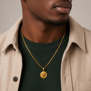 Op de afbeelding: Een goudkleurige metalen ketting met een ronde hanger voorzien van een gedetailleerd reliëf van een klassiek gezicht. De ketting rust op een donkergroene geribbelde trui die onder een lichtbeige overhemd met knopen wordt gedragen.
