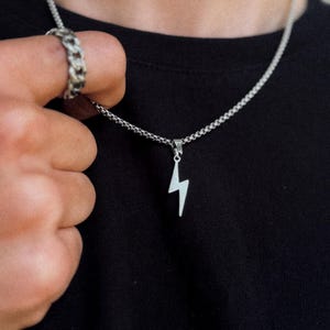 Könnte beinhalten: Silberfarbene Halskette mit einem Blitz-Anhänger. Die Halskette wird vor einem schwarzen T-Shirt getragen. Ein silberner Ring ist am Finger der Person zu sehen. Die Kette hat ein strukturiertes Design.