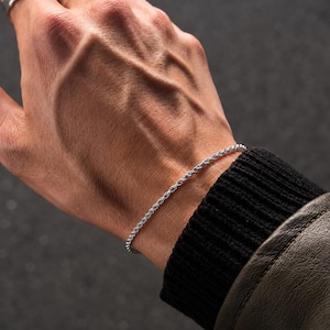 Pode incluir: Uma pulseira de corrente de corda prateada é usada no pulso. A pulseira é feita de pequenas elos bem tecidos. A pessoa está usando um suéter preto e uma jaqueta de couro marrom. O fundo é cinza escuro.
