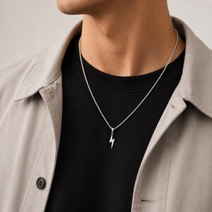 Op de afbeelding: Een zilverkleurige ketting met een kleine, minimalistische bliksemschicht-hanger aan een dunne, getextureerde touwketting. De hanger hangt gecentreerd op een zwart shirt dat onder een lichtbeige overhemd met kraag wordt gedragen.