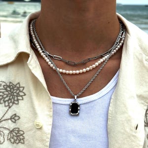 Op de afbeelding: Een gelaagde ketting met een zilveren ketting, een parelstreng en een hanger met een zwarte steen. De hanger is rechthoekig met een getextureerde rand.