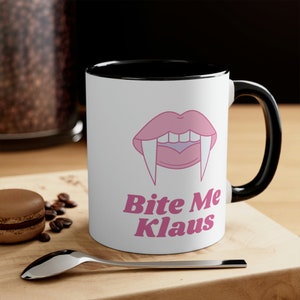 May include: White coffee mug with a black rim and handle. The mug has a pink cartoon vampire mouth with fangs and the text "Bite Me Klaus" in pink.