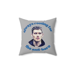 May include: A gray throw pillow with a blue circle around a photo of a man with the text "always rooting for the anti-hero" in blue.