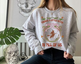 May include: A light gray sweatshirt with long sleeves featuring the text "THERE'S A LITTLE WITCH IN ALL OF US". The design includes a witch on a broomstick in a decorative frame. The sweatshirt is a casual, comfortable style.
