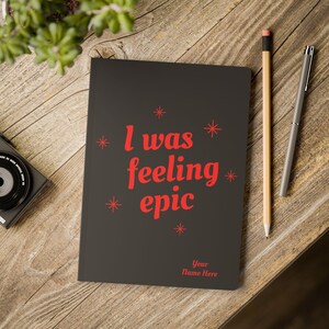 May include: A black notebook with the text "I was feeling epic" in red, along with red starburst designs. A pencil and pen are next to the notebook on a wooden surface. The notebook also has the text "Your Name Here".