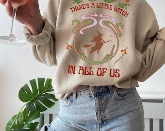 May include: Beige sweatshirt with the text "THERE'S A LITTLE WITCH IN ALL OF US" in red and green. The design includes a witch on a broomstick. The person is wearing ripped blue jeans and holding a glass of wine.