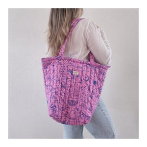 May include: A quilted tote bag in bright pink featuring a grid pattern with blue line art illustrations of celestial bodies, moths, snakes, and plants. The bag has two shoulder straps and a small rectangular label with the text Letie.