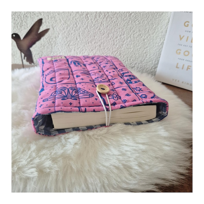 May include: A quilted fabric book sleeve in bright pink with a dark blue celestial print featuring moons, stars, and moths. The sleeve has a light-colored wooden button closure with an elastic cord and a blue and white gingham interior lining.