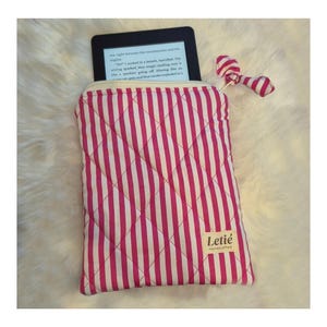 May include: A quilted fabric sleeve with a pink and white vertical striped pattern containing an e-reader. The sleeve features a small fabric bow attached to the top corner and a rectangular beige label with the text Letié Handcrafted.