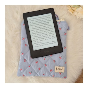 May include: A black e-reader with a screen displaying text resting on a light blue quilted fabric sleeve. The sleeve features a repeating pattern of small red cherries and a white rectangular tag with the text Letié Handcrafted.