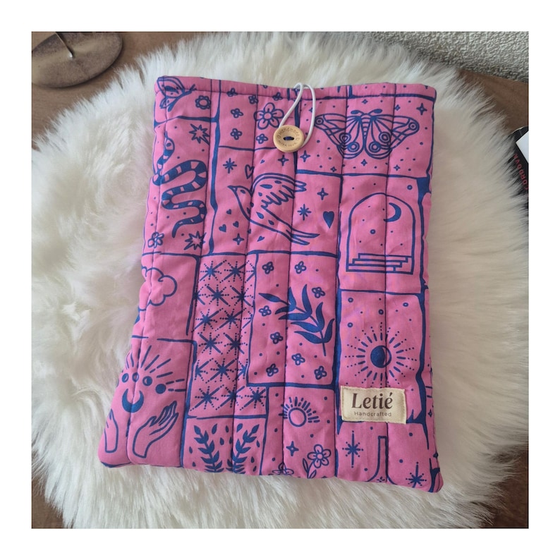 May include: A quilted fabric sleeve in bright pink with a repeating dark blue graphic print featuring butterflies, snakes, birds, celestial symbols, and hands. It includes a wooden button closure with a white cord and a Leti&eacute; Handcrafted label.