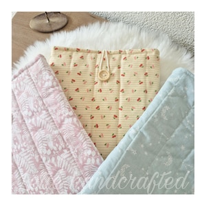 May include: Three quilted fabric pouches featuring distinct patterns: a cream pouch with red cherry prints and a wooden button closure, a pink pouch with white fern leaf patterns, and a light blue pouch with white moon and star motifs.