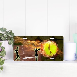May include: A yellow softball with black stitching is engulfed in flames. Two silhouettes of softball players are on a baseball field. The image is on a black background with a brown border.