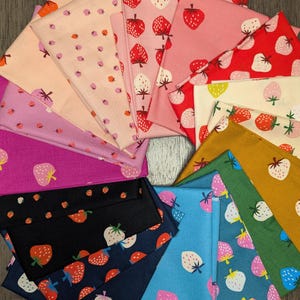 May include: A collection of folded fabric squares in various colors, each featuring a strawberry pattern. Colors include pink, red, black, blue, and green. The strawberries have red, white, and pink details. The fabric is likely cotton or a similar material.