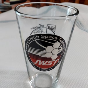May include: A clear glass with a black and white logo for the James Webb Space Telescope. The logo includes the text "James Webb Space Telescope", "JWST", "NASA", "ESA", and "CSA".