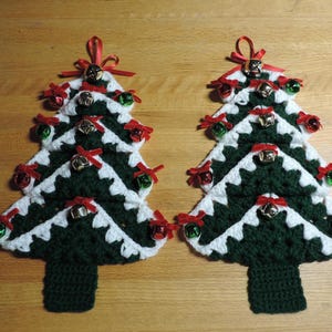 May include: Two crocheted Christmas tree ornaments featuring alternating dark green and white yarn layers. Each tree is decorated with small red ribbon bows and metallic jingle bells in red, green, and silver colors, with a red ribbon loop at the top.