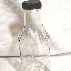 May include: A clear glass bottle with a black lid. The bottle has a ridged design on the sides and a rounded base. The bottle is empty and sits on a white textured surface. The bottle is suitable for storing liquids.