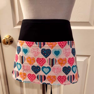 May include: A black and white heart-patterned apron with a black waistband and a single pocket. The apron is tied at the waist with black strings.