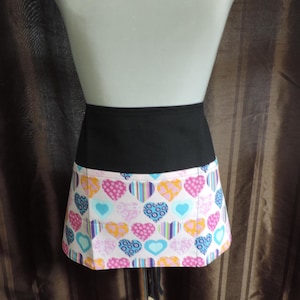 May include: A waist apron featuring a solid black upper band and a lower section patterned with multicolored hearts in shades of pink, blue, orange, and purple. The heart designs include various textures like stripes, dots, and swirls on a light background.