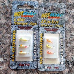 May include: Two packages of fishing lures, one labeled "Hot Bite Lures Little Stinger Jigs" and the other labeled "Hot Bite Lures Darter Micro Jigs". Both packages feature a clear plastic window showing three lures each. The lures are yellow, orange, and pink with black eyes.