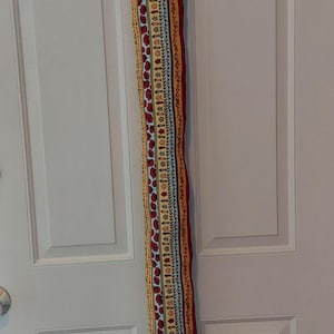 May include: A long, rectangular fabric door draft stopper with a colorful pattern of red, yellow, and blue designs. The stopper is hanging on a white door with a red ribbon.
