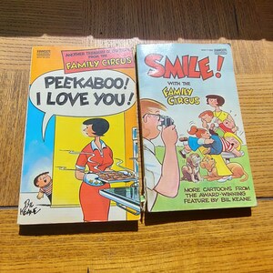 May include: Two vintage Family Circus comic books. One cover reads "Peekaboo! I Love You!" with a cartoon of a woman in a red dress. The other cover says "Smile!" with a cartoon of a family. Both books are on a wooden surface.
