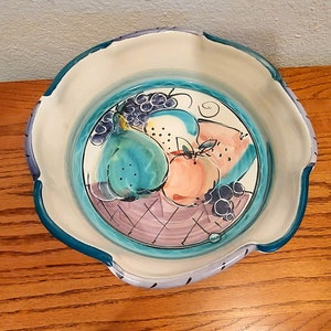 May include: A white ceramic bowl with a blue, green, and purple fruit design. The bowl has a scalloped edge and is perfect for serving fruit or snacks.