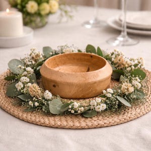 May include: A light brown wooden bowl sits on a woven placemat, surrounded by a wreath of white flowers and green leaves. The bowl is the focal point, with a smooth, natural finish. The arrangement suggests a rustic, natural theme.