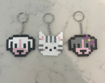 May include: Three keyrings with pixel art animal faces: a dog, a cat, and another dog. The keyrings are made of colourful beads, with silver key rings and chains. The dog keyrings have pink and black details, while the cat has grey stripes.