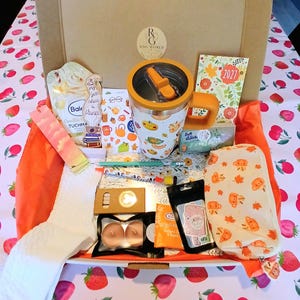 Könnte beinhalten: Eine Geschenkschachtel aus Pappe, gefüllt mit verschiedenen Artikeln, darunter ein weißer Reisebecher mit orangefarbenem Deckel und herbstlichen Motiven, ein Federmäppchen mit Kürbismuster, ein Kalender für 2027, weiße Socken und Teelichter.