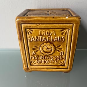 May include: A square, ceramic container in a warm, golden-brown hue. The front features the text "FROM SANTA CLAUS" above a circular emblem and the words "A. MATTHEWS SONS BROOKLYN". The top has a floral design.