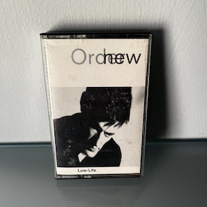 May include: A cassette tape with a black and white image of a person on the cover. The text "Orchew" is at the top, and "Low-Life" is at the bottom. The cassette is in a clear plastic case.