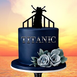 May include: A blue cake with a white base, decorated with a silhouette of two people on a railing, the word "TITANIC" in silver lettering, and blue roses.